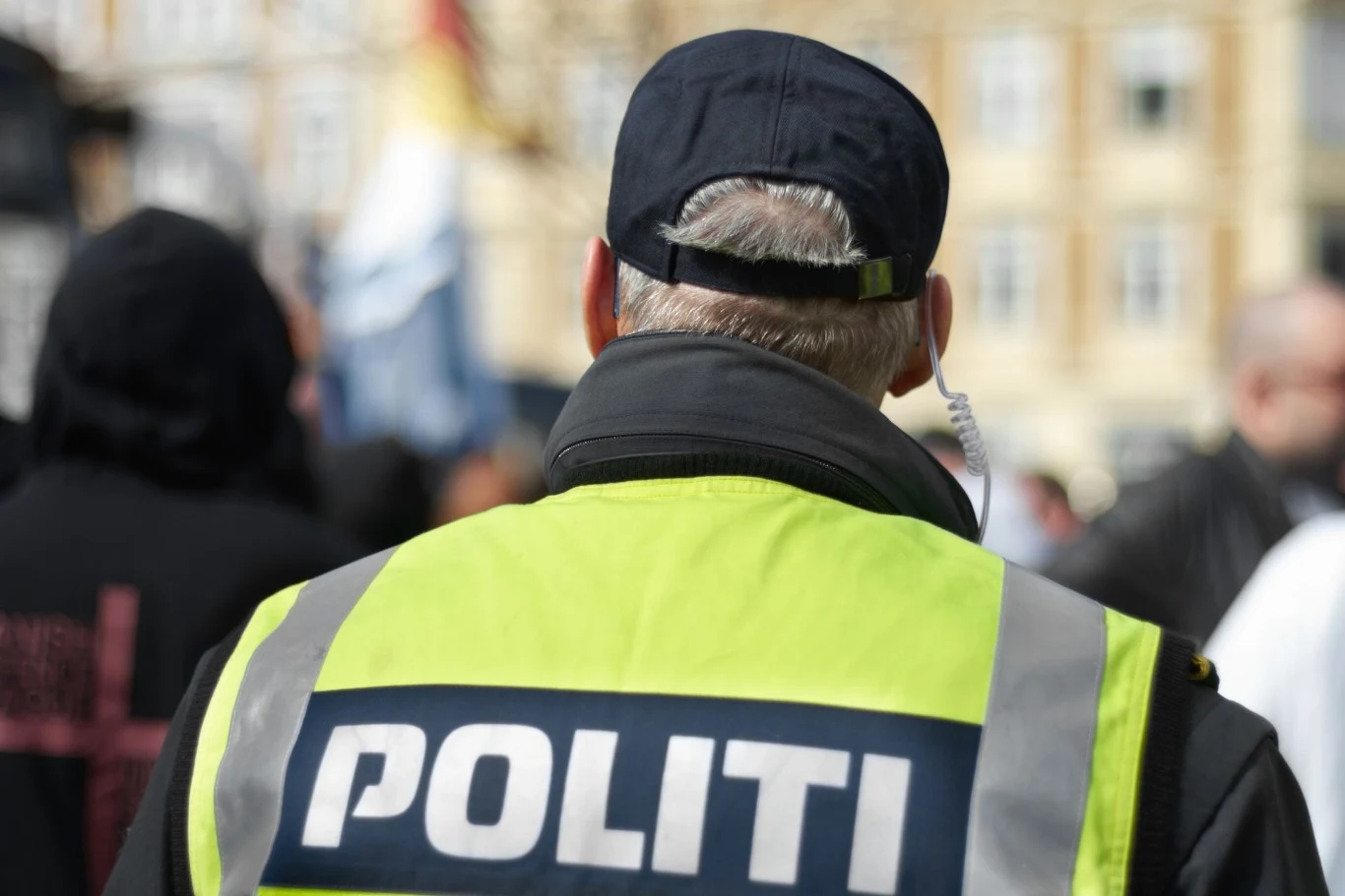 Protesty często zamieniały się w potyczki z policją. Policjant w jaskrawej kamizelce z napisem 'POLITI' patrzący w stronę tłumu. Tło zamazane, co wskazuje na sytuację miejską, prawdopodobnie podczas wydarzenia publicznego lub manifestacji.