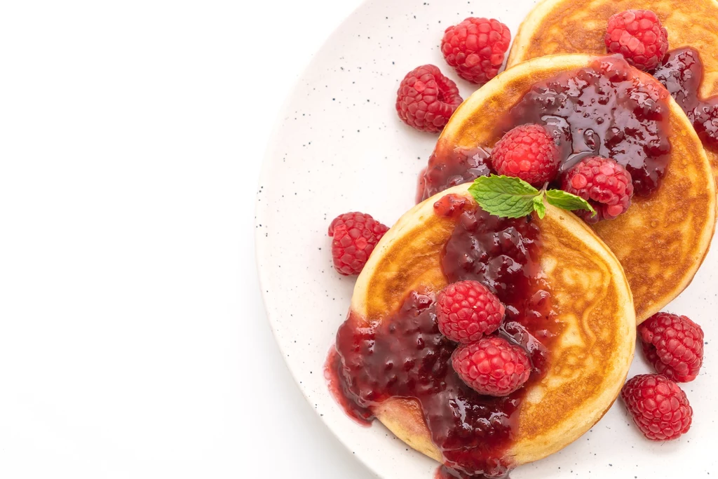 Puszyste pancakes są jednocześnie kwaśne i słodkie, co gwarantuje niesamowite doznania smakowe Naleśniki z malinowym sosem i świeżymi malinami na białym talerzu, ułożone na białym tle.