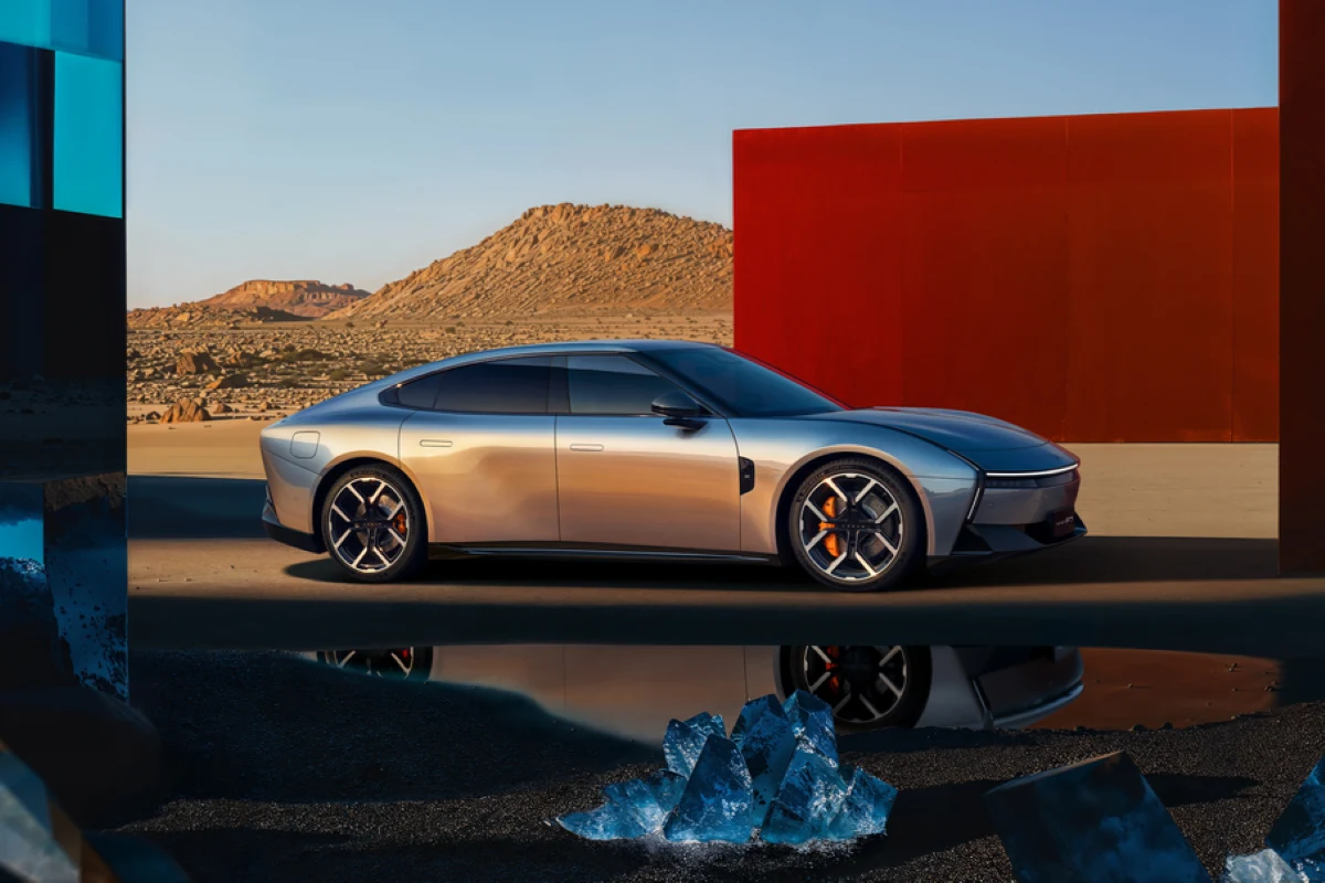 Luksusowy, srebrny samochód elektryczny stoi na pustynnym tle w otoczeniu nowoczesnych, kolorowych instalacji. Refleksy na karoserii odbijają błyszczące, szkliste kamienie na ziemi. W tle widać skaliste wzgórza.