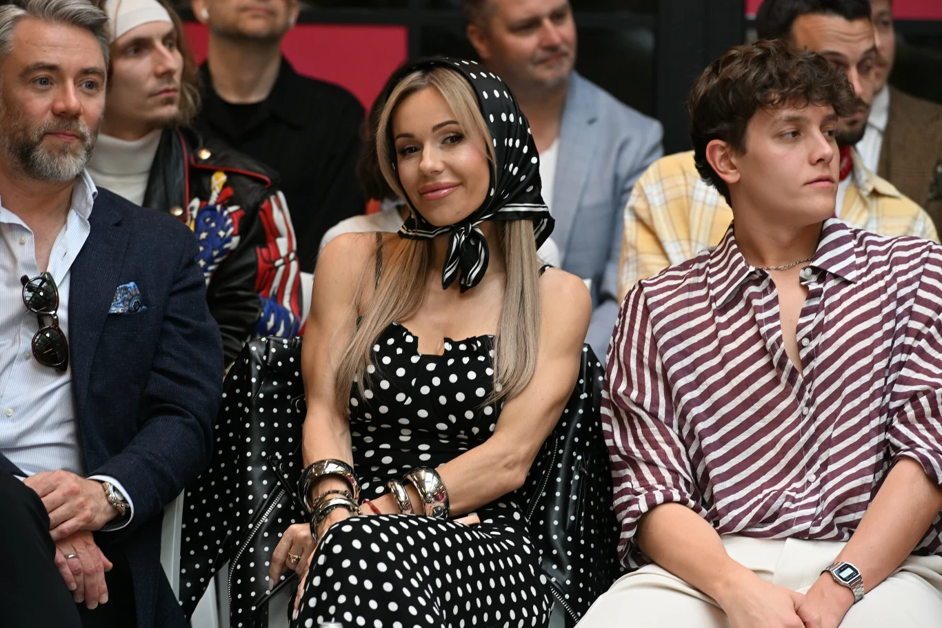 Grupa osób siedzących w rzędzie, uśmiechnięta kobieta w sukience w grochy i chustce na głowie pośród mężczyzn ubranych w eleganckie i codzienne ubrania, prawdopodobnie uczestnicząca w jakimś wydarzeniu.