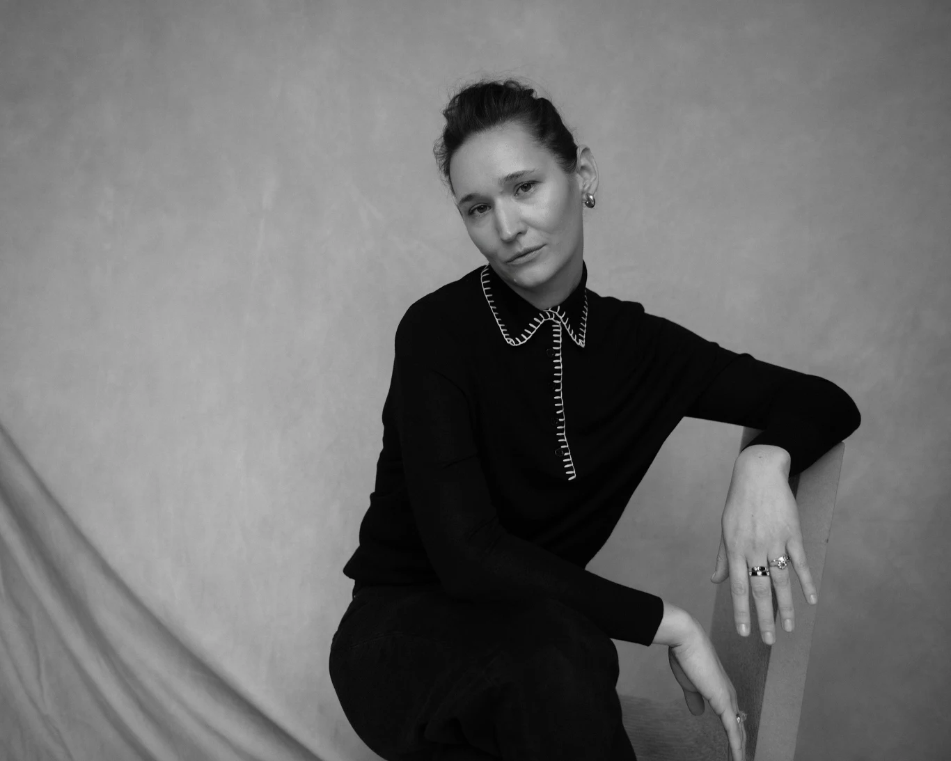 Czarno-biała fotografia przedstawiająca osobę siedzącą w luźnej pozie. Ubrana jest w ciemny sweter z ozdobnym kołnierzem i nosi kilka pierścionków. Tło jest neutralne, co podkreśla sylwetkę postaci.