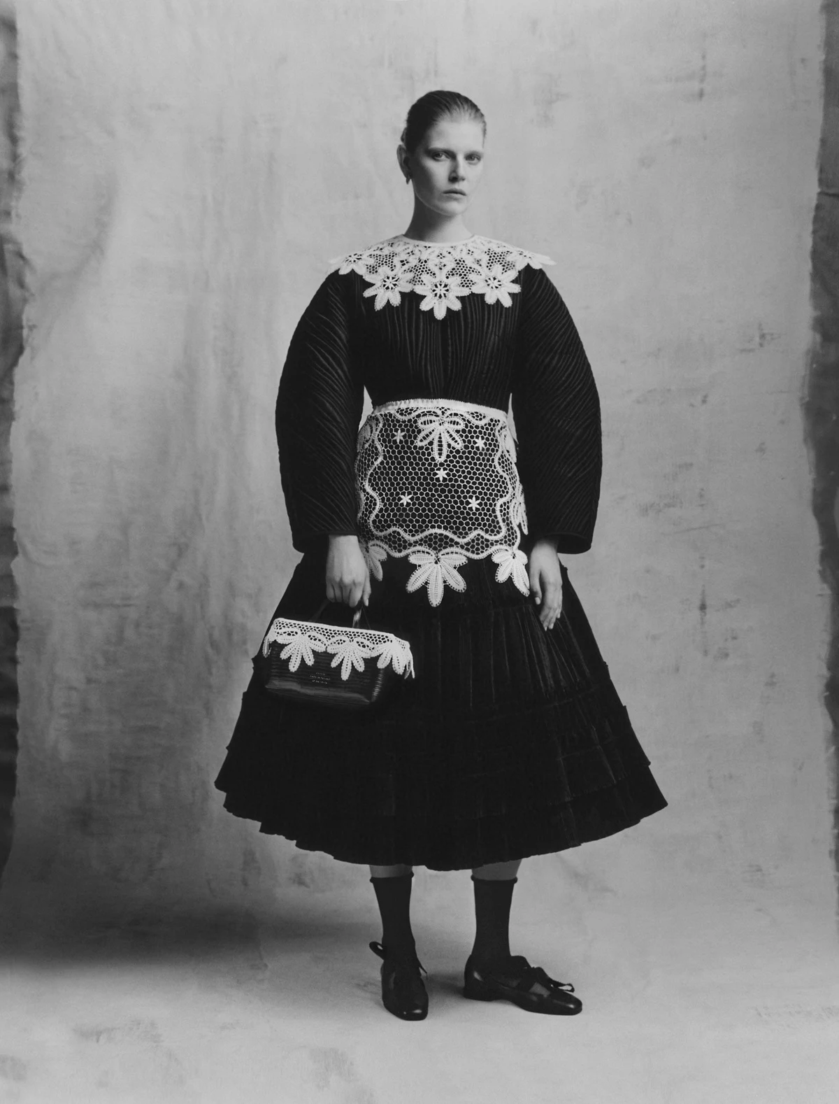 Nowa kolekcja Zofii Chylak to hołd dla tradycji rzemieślniczej z Bobowej. W roli głównej: koronka (zdj. pochodzi z kampanii Chylak Heritage) Osoba w ciemnej sukni z koronkowymi wykończeniami. Suknia ma szerokie rękawy i ozdobne detale na dekolcie oraz kieszeni. Kobieta trzyma torebkę z podobnym wzorem.