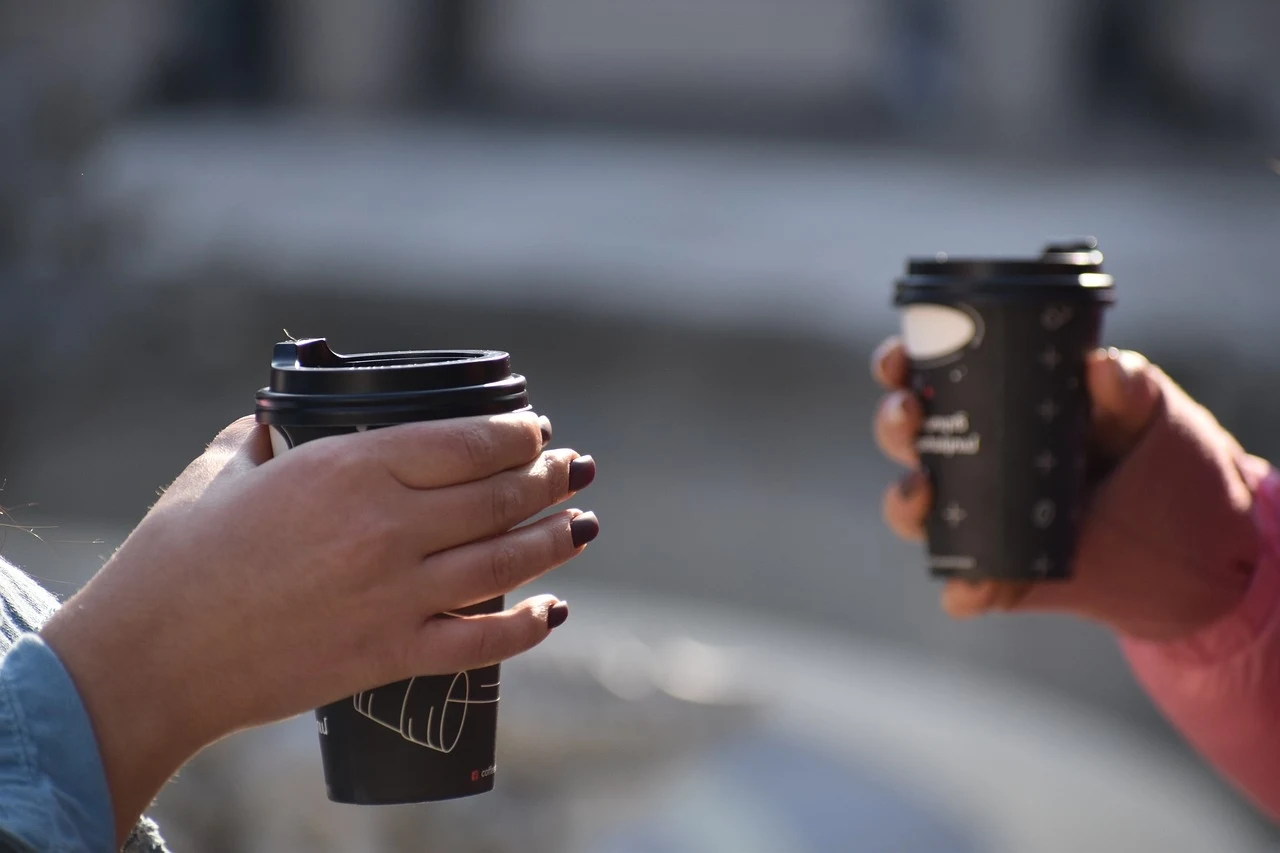Dwie osoby trzymające kubki z kawą na wynos, jedna z nich ma paznokcie pomalowane na ciemny kolor. Scena rozgrywa się na zewnątrz, w słoneczny dzień.