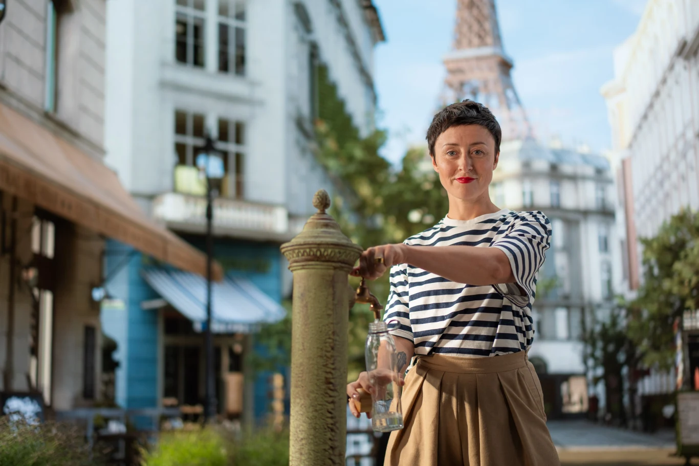 Kobieta nalewająca wodę z miejskiego kranu, ubrana w pasiastą bluzkę i spódnicę, stoi pośrodku ulicy w otoczeniu paryskiej architektury, w tle widoczna wieża Eiffla.