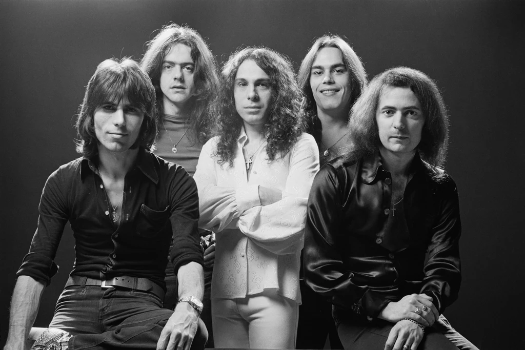 Rainbow w 1976 r. - w środku Ronnie James Dio, pierwszy z prawej Ritchie Blackmore Grupa pięciu mężczyzn, ustawiona w dwóch rzędach, o długich włosach, ubrana w ciemne i jasne koszule, na neutralnym tle. Wyraz ich twarzy jest poważny, sylwetki subtelnie oświetlone studyjnym światłem.