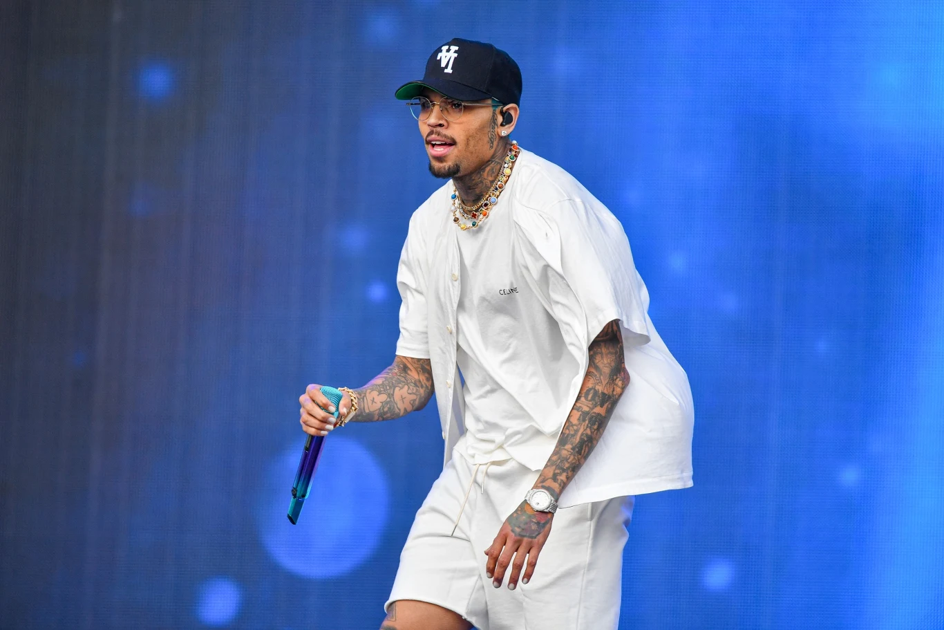 Chris Brown został zatrzymany Mężczyzna ubrany w białą koszulkę i szorty, z tatuażami na ramionach, trzyma mikrofon na scenie. Ma na sobie czapkę z daszkiem, a w tle jest jasnoniebieska scena.