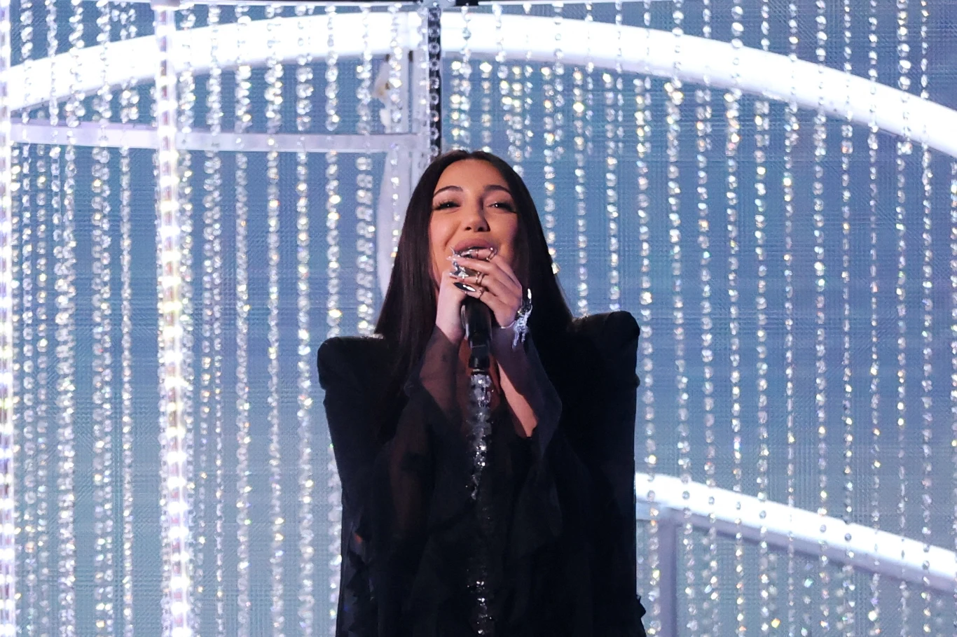 Juwal Rafael jest reprezentantką Izraela na Eurowizji 2025 w Bazylei Kobieta w eleganckiej czarnej kreacji śpiewa na scenie, trzymając mikrofon. W tle znajduje się efektowna dekoracja złożona z błyszczących, świetlnych elementów.