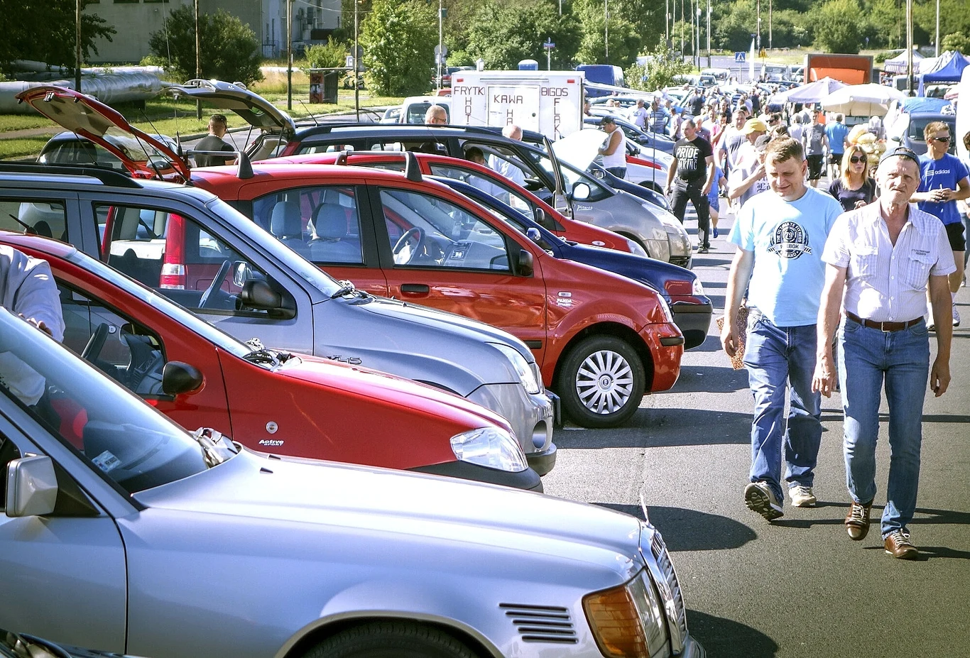 Handlarz wyjaśnia, skąd się wziął ogłoszeniowy slang. To spadek po czasach płatnego roamingu Targ motoryzacyjny z rzędem zaparkowanych samochodów osobowych. Po lewej stronie kilka otwartych bagażników, po prawej ludzie spacerujący i oglądający auta. W tle stoiska wystawców i drzewa.