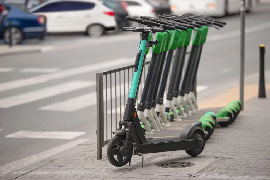 Zarejestrowanie hulajnogi elektrycznej, będącej motorowerem może być niemożliwe. Szereg hulajnóg elektrycznych zaparkowanych na chodniku przy ruchliwej ulicy. Można zauważyć kilka samochodów w tle oraz przejście dla pieszych. Hulajnogi mają zielono-czarne barwy.