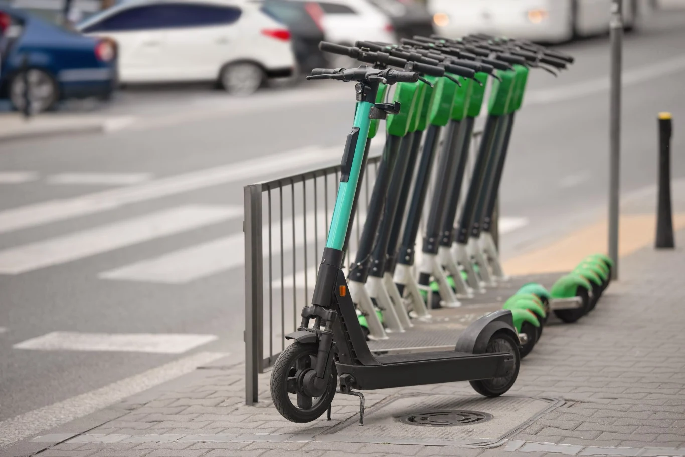Zarejestrowanie hulajnogi elektrycznej, będącej motorowerem może być niemożliwe. Szereg hulajnóg elektrycznych zaparkowanych na chodniku przy ruchliwej ulicy. Można zauważyć kilka samochodów w tle oraz przejście dla pieszych. Hulajnogi mają zielono-czarne barwy.