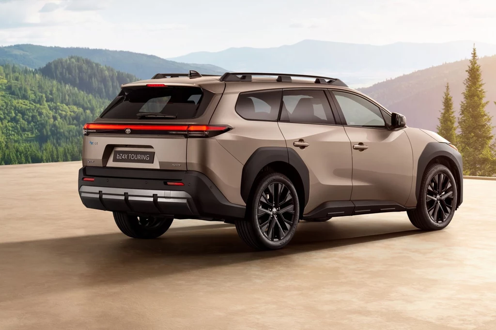 Toyota bZ4X Touring będzie dostępna w dwóch wariantach mocy: 224 KM (napęd na przód) i 380 KM (napęd na cztery koła). Samochód typu SUV w nowoczesnym designie zaparkowany na tle malowniczego krajobrazu górskiego. Pojazd w kolorze beżowym z ciemnymi szybami i sportowymi felgami. W tle widoczne zielone wzgórza i drzewa.