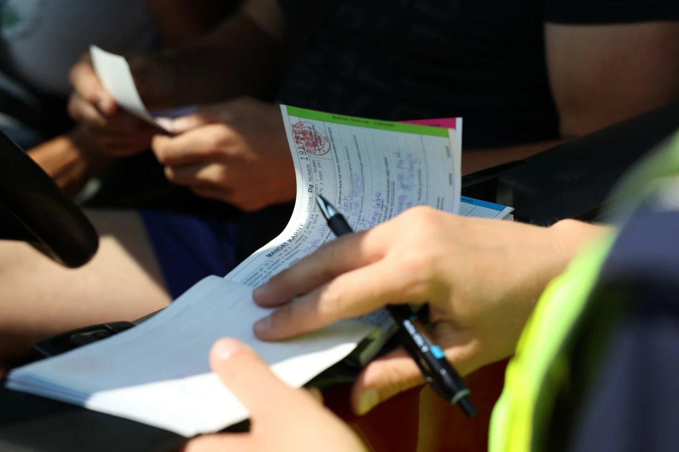 Ręce trzymające różne dokumenty i długopisy, jedna z osób pisze notatkę. Jasne światło podkreśla szczegóły na dokumentach. Sytuacja sugeruje załatwianie formalności lub wykonywanie pracy związanej z dokumentacją.