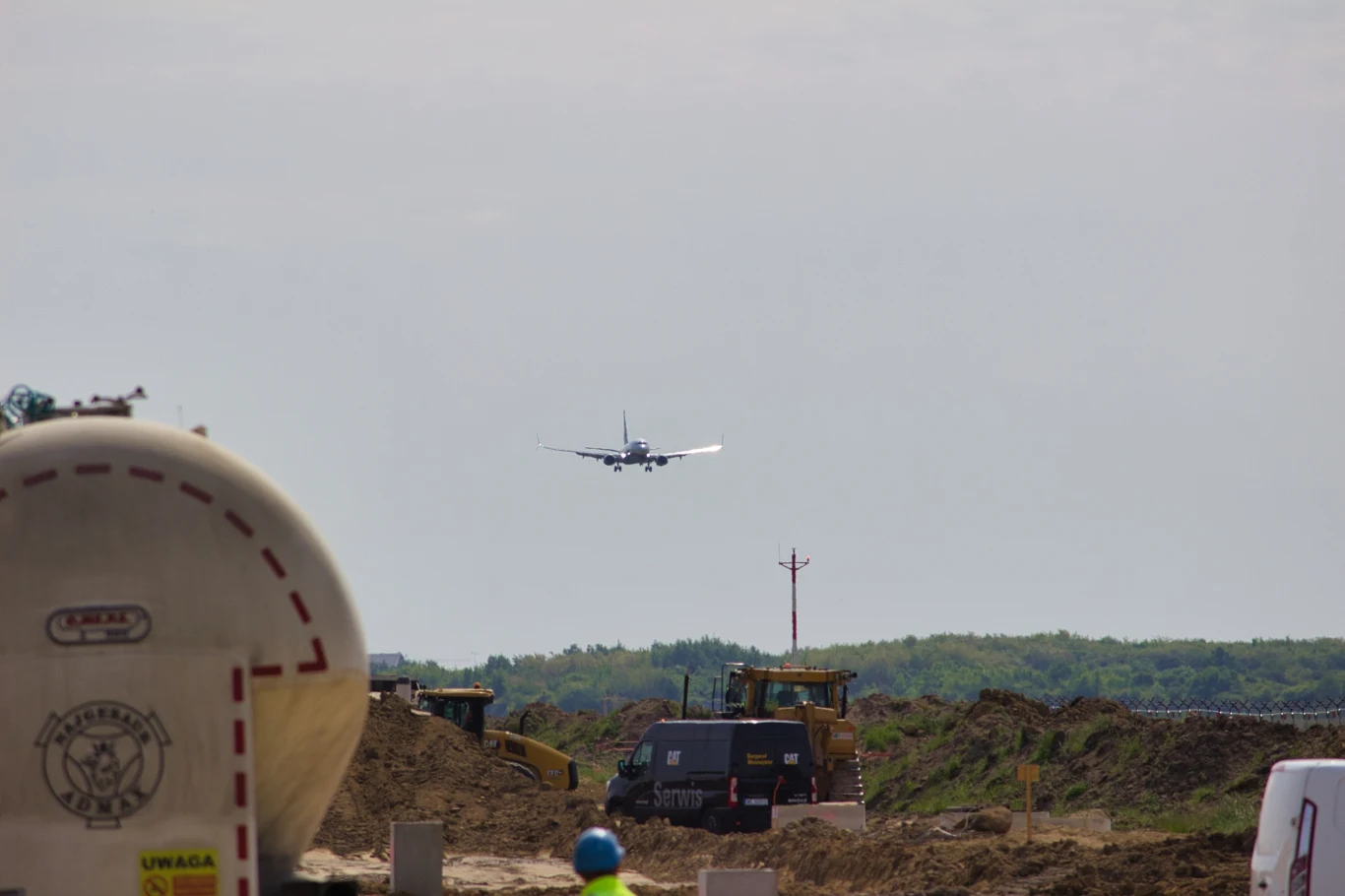 Prace budowlane na wrocławskim lotnisku obecnie trwają przy aktywnym ruchu lotniczym. Samolot w trakcie lądowania na lotnisku, wokół trwają działania budowlane, na pierwszym planie widoczny zbiornik oraz maszyny budowlane. Niebo częściowo zachmurzone, co tworzy spokojną scenerię.