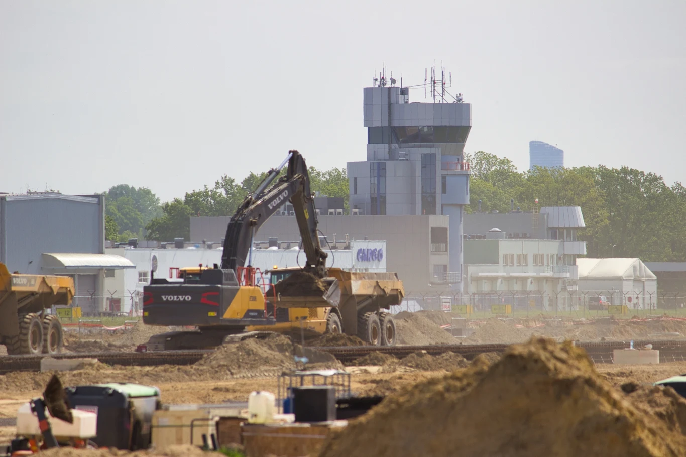 Lotnisko we Wrocławiu zostanie zamknięte na 40 dni - od 26 października do 4 grudnia 2025. Plac budowy z koparką i ciężarówkami w tle, na którym znajduje się budynek kontrolny lotniska. Wokół rozciąga się teren otoczony niską roślinnością, a na pierwszym planie widoczne są usypane hałdy ziemi.