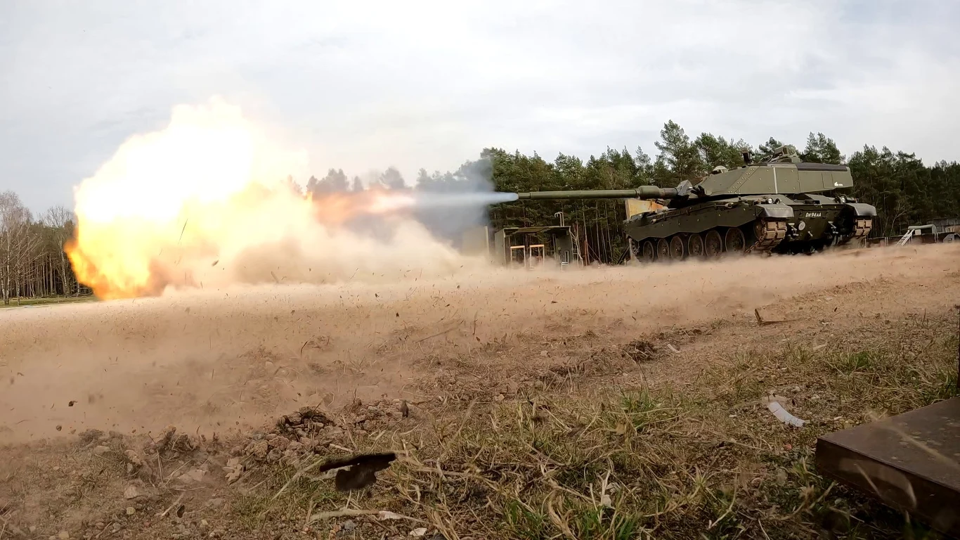 Prototyp czołgu Challenger 3 podczas testów poligonowych Czołg strzelający w kierunku nieba, wystrzał generuje jasny płomień i kłęby dymu. Otwarta przestrzeń, w tle widoczne drzewa.