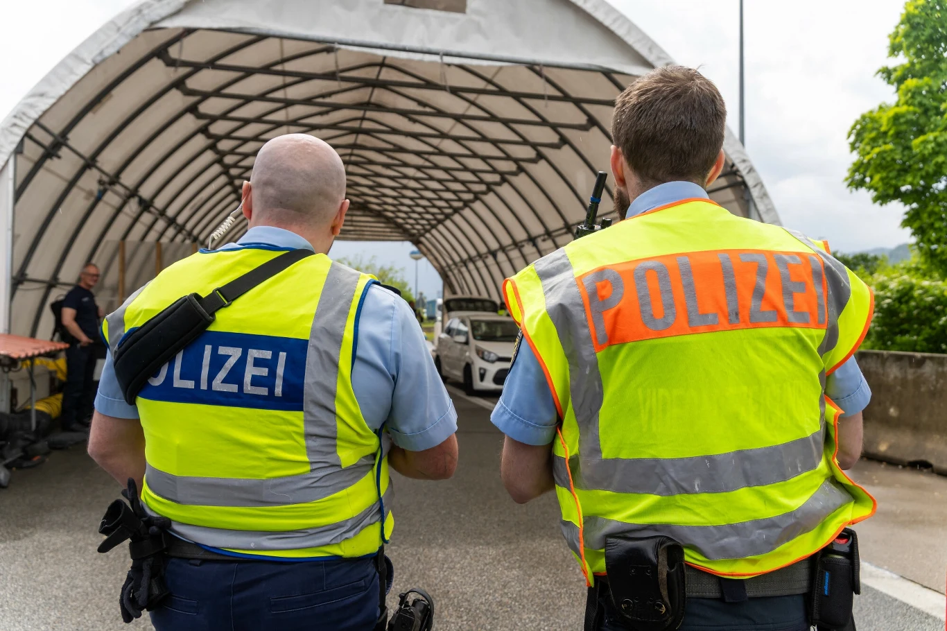 Wyjeżdżasz do Niemiec w czerwcu? Szykuj się na kontrole i utrudnienia. Dwaj policjanci w kamizelkach odblaskowych z napisem 'Polizei' stoją przed tymczasowym zadaszonym stanowiskiem kontroli drogowej. Mężczyźni są odwróceni plecami do obiektywu.