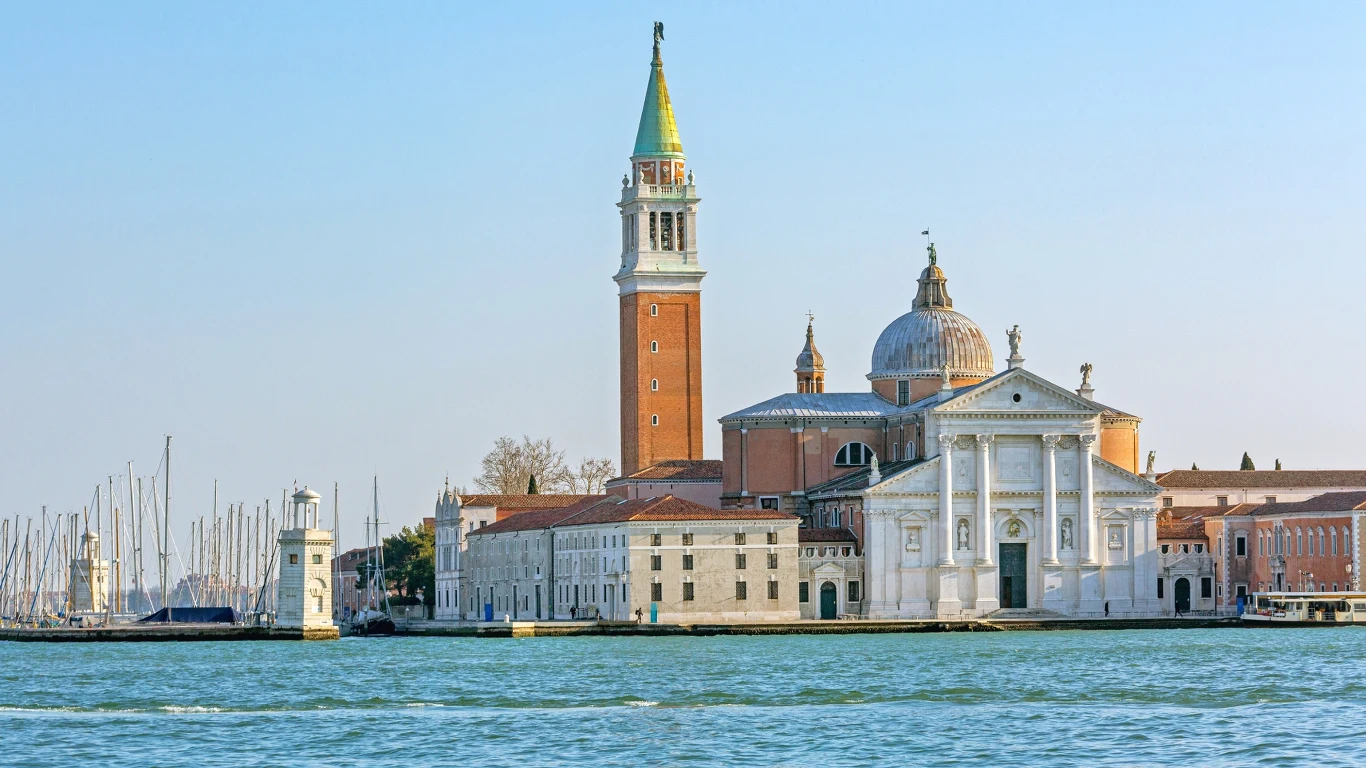 Malowniczy widok na kościół San Giorgio Maggiore w Wenecji, z charakterystyczną wieżą i kopułą. W tle widać spokojną wodę i cumujące łodzie, a architektura zachwyca renesansowym stylem.