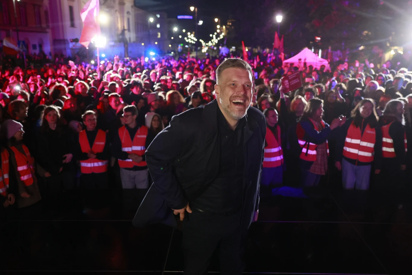 Adrian Zandberg podczas wiecu w Warszawie, wybory prezydenckie 2025 Mężczyzna uśmiechający się na pierwszym planie w otoczeniu tłumu ludzi ubranych w czerwone kamizelki. W tle widać kolorowe światła i elementy architektury miasta nocą.