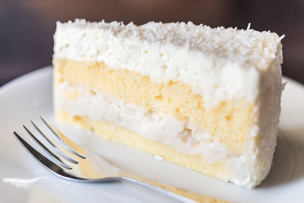 Pyszny tort Raffaello Kawałek kremowego ciasta z jasnym biszkoptem, przełożonego warstwą delikatnego kremu i pokrytego obficie wiórkami kokosowymi, na białym talerzu z widelcem obok. Ciasto wygląda na puszyste i apetyczne.