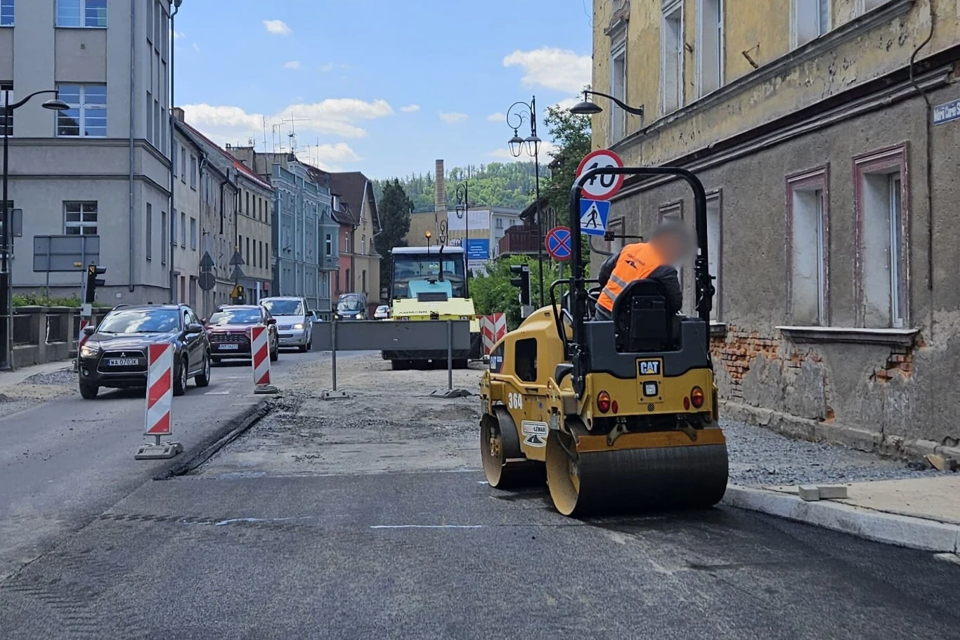 Pracownik budowlany obsługujący walec drogowy na ulicy w trakcie remontu. Obok widoczne są pachołki ostrzegawcze i znaki drogowe. W tle znajdują się budynki i zaparkowane samochody.