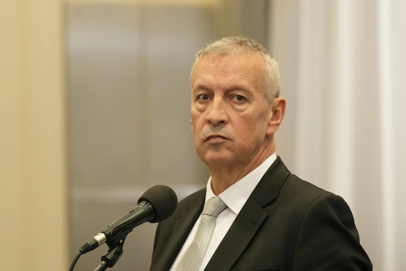 Rzecznik szefa MSWiA Jacek Dobrzyński Mężczyzna w garniturze stoi przed mikrofonem, spoglądając w stronę obiektywu. Jego postawa jest poważna, a tło jest neutralne.