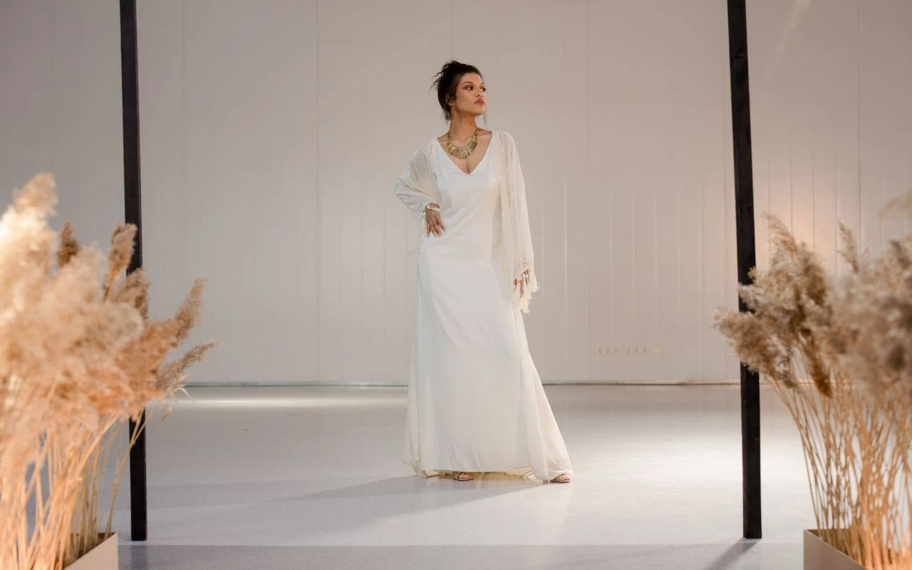 Kobieta w białej, długiej sukni stoi na jasnym tle pomiędzy dekoracjami z suszonych traw. Jej wyraz twarzy jest spokojny, a dłonie spoczywają luźno na biodrach. Stylizacja ma elegancki charakter, a biżuteria dodaje jej szyku.