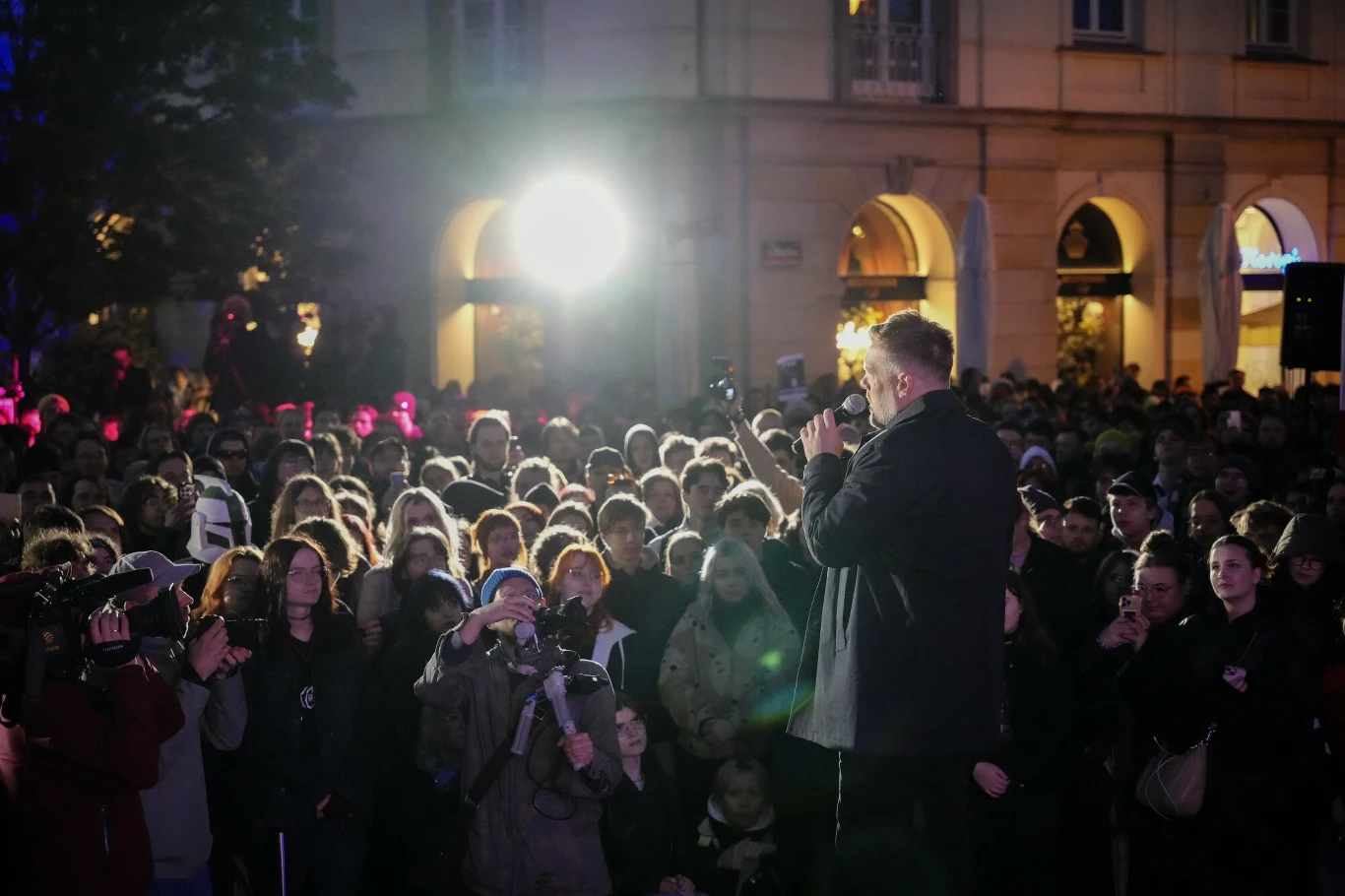 Mężczyzna przemawiający do tłumu podczas wydarzenia na otwartej przestrzeni, wieczorową porą, w miejskim otoczeniu. W tle oświetlone budynki i jasne reflektory.