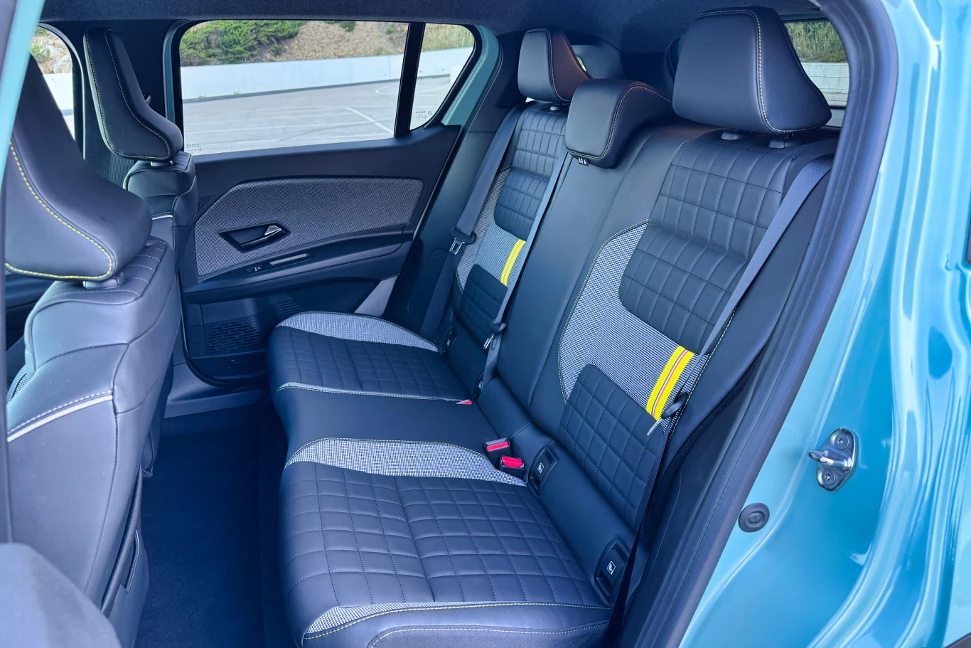 Wnętrze samochodu z tylnymi siedzeniami pokrytymi czarną tapicerką o wzorze kratki. Siedzenia wyposażone w pasy bezpieczeństwa w kolorze żółtym. W tle boczne okna i drzwi o modernistycznym designie.