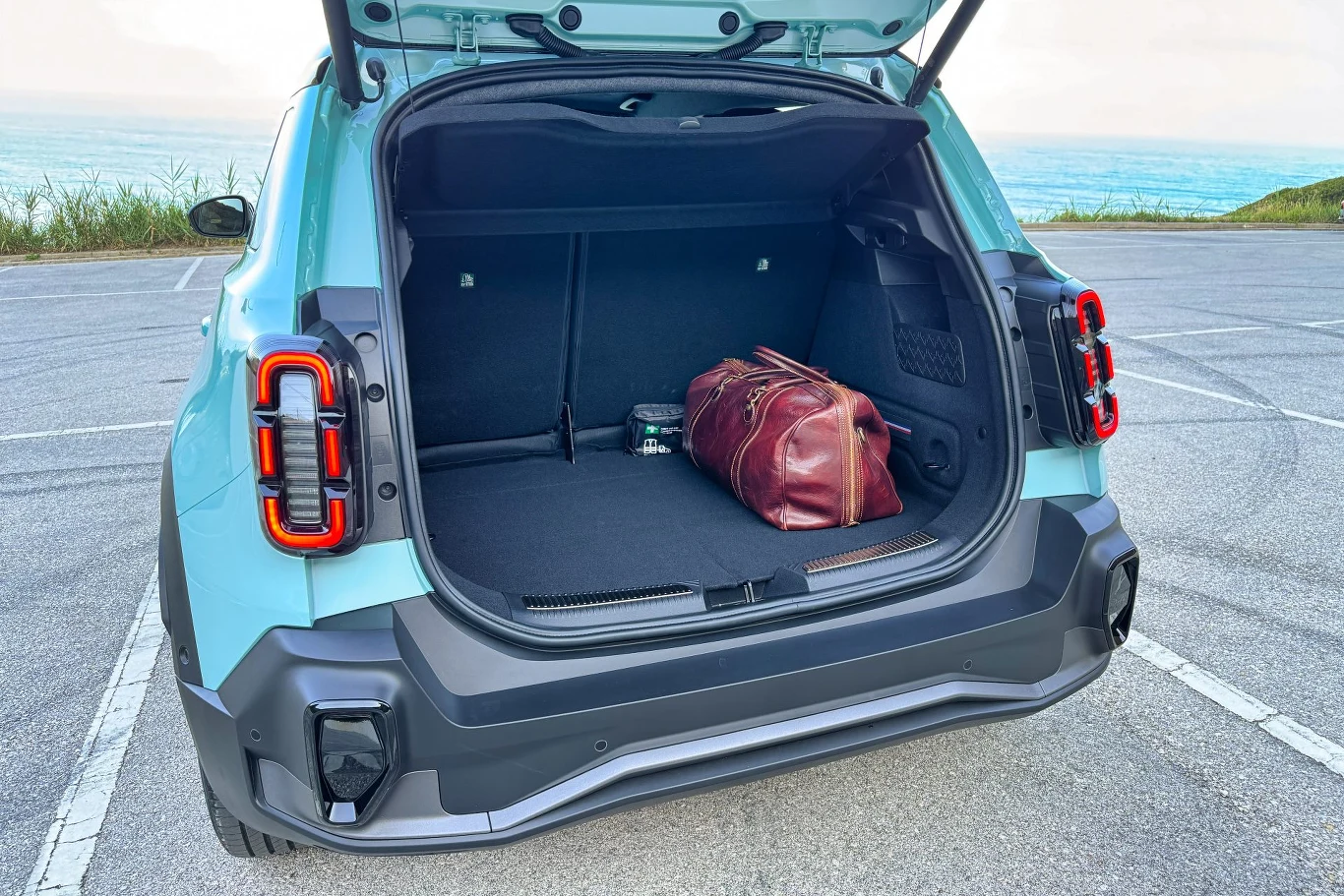 Otwarty bagażnik turkusowego SUV-a zaparkowanego blisko wybrzeża. W środku umieszczona jest skórzana torba podróżna.