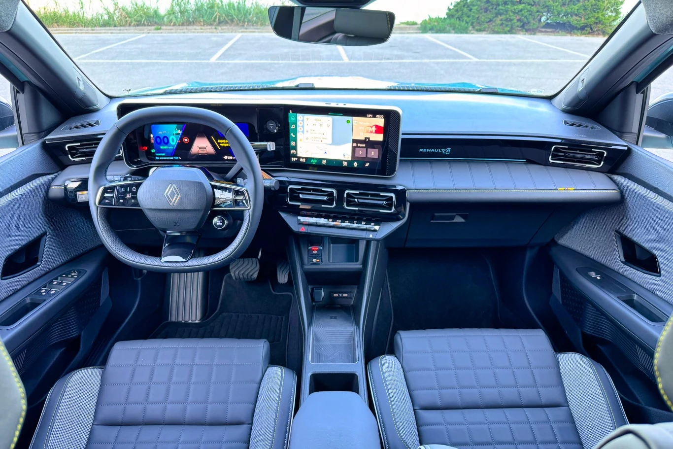 Wnętrze nowoczesnego samochodu z widocznym kierownicą z logo Renault, nowoczesnymi wyświetlaczami i eleganckimi siedzeniami. Deska rozdzielcza z ekranem dotykowym, a w tle widoczny parking.