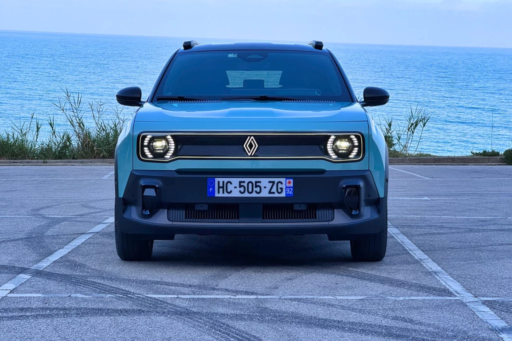 Z przodu podświetlone jest nie tylko logo, ale i cała ramka wokół "grilla" Samochód marki Renault zaparkowany nad brzegiem morza, w tle rozciąga się niebieski horyzont. Przód pojazdu z charakterystycznym logo i światłami, ustawiony na wprost aparatu. Otoczenie tworzy spokojny, nadmorski klimat.