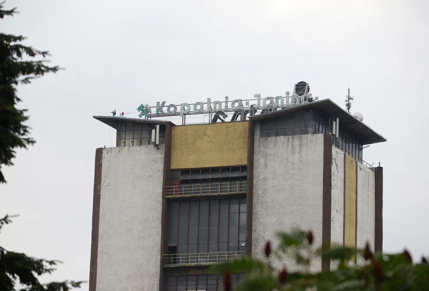 Wstrząs w kopalni "Janina" w Libiążu Wieżowiec z logo Kopalni Janina na dachu, otoczony zielenią. Biała fasada z brązowymi wzmocnieniami konstrukcyjnymi oraz dużymi oknami na froncie budynku. Chmury na niebie w tle.