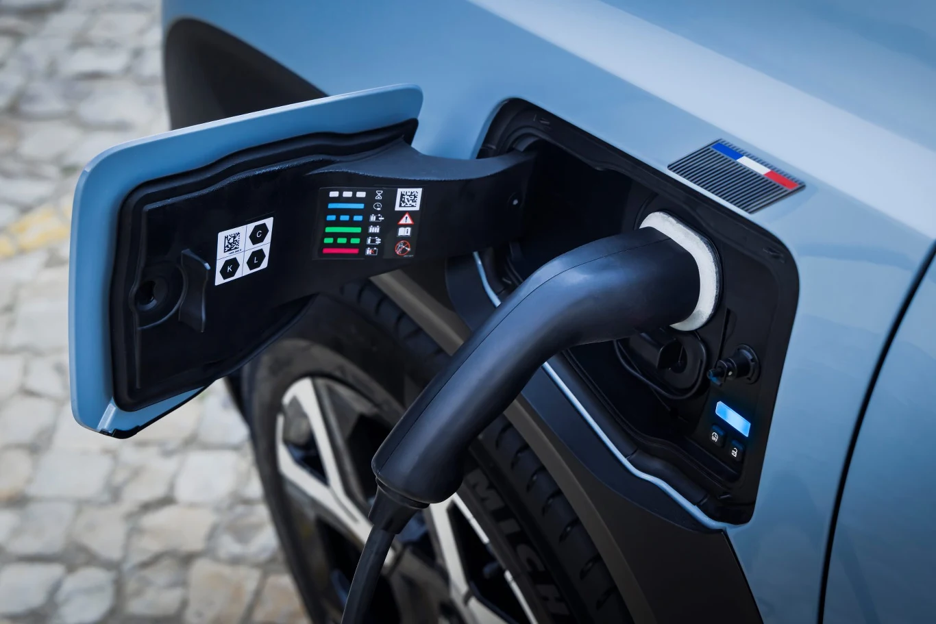 Zbliżenie na gniazdo ładowania samochodu elektrycznego, w którym wpięta jest wtyczka ładowarki. Obok znajdują się naklejki z informacjami o ładowaniu oraz flaga narodowa. Karoseria samochodu ma niebieski kolor.