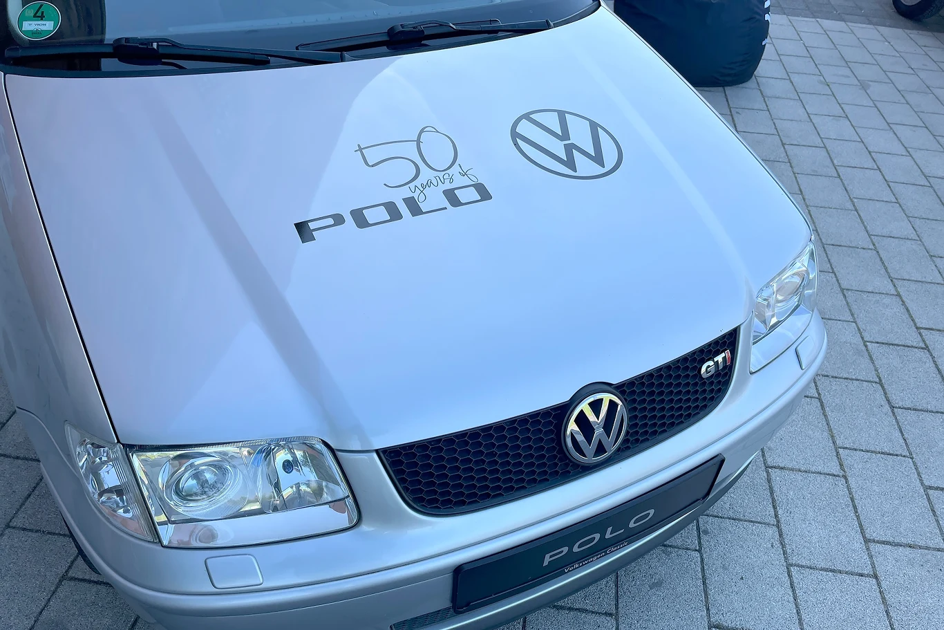 Volkswagen Polo ma już 50 lat. Sześć generacji i dziesiątki wariantów Srebrny samochód Volkswagen Polo z widocznymi logotypami VW i napisami na masce, stoi na kostce brukowej. Na masce widoczna jest liczba '50', co może wskazywać na rocznicę modelu.