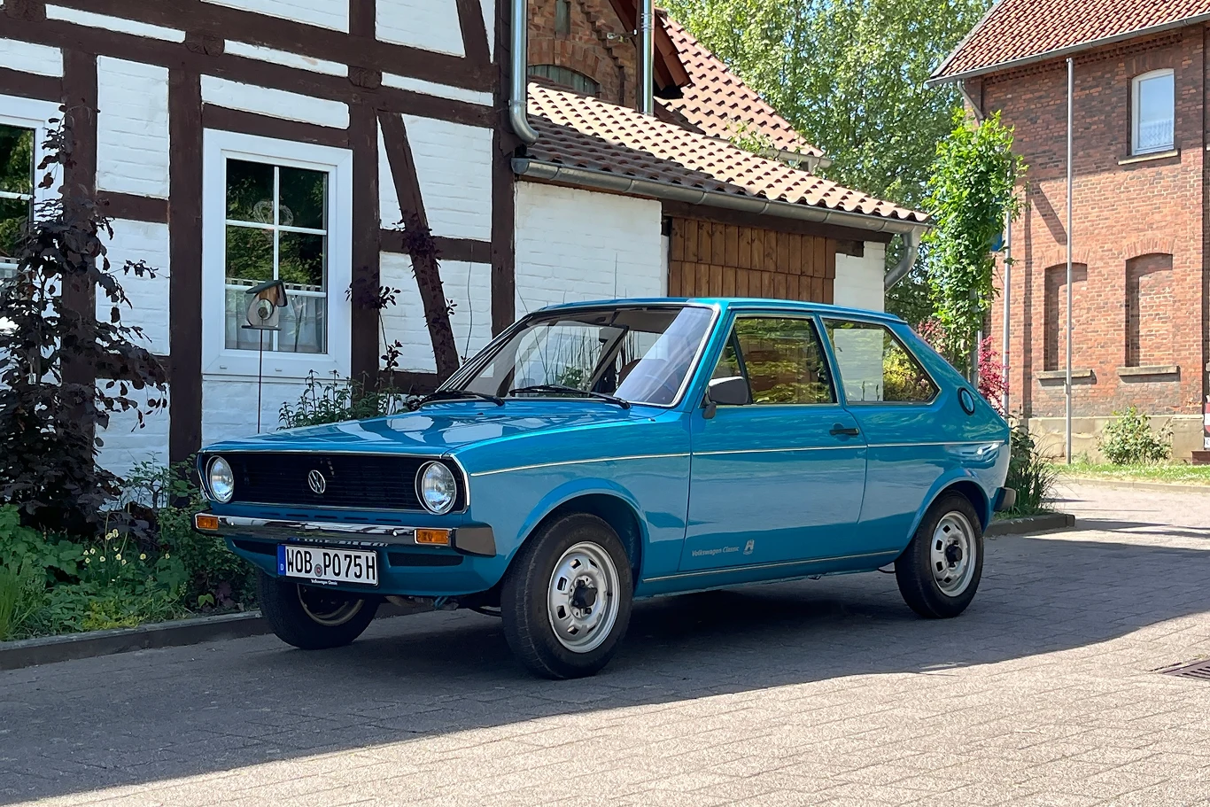 Pierwszy Volkswagen Polo to budżetowa odpowiedź na Audi 50. Klasyczny, niebieski samochód zaparkowany obok tradycyjnego domu z muru pruskiego, na tle spokojnej, zielonej okolicy.