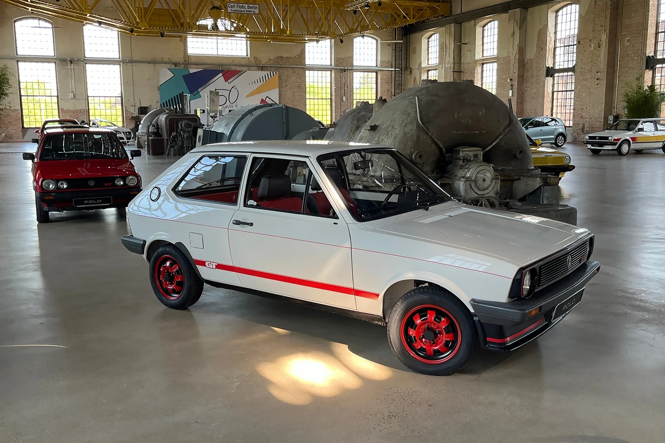Prawdziwą furorę zrobił limitowany model Polo Coupé GT40 wyposażony w 1,3-litrowy silnik z doładowaniem. Samochód osobowy w stylu retro stoi w dużej, oświetlonej hali przemysłowej. W tle znajdują się inne klasyczne pojazdy oraz ciężkie, przemysłowe maszyny. Główna uwaga skupia się na białym aucie z czerwonymi felgami i dekoracyjnymi paskami.