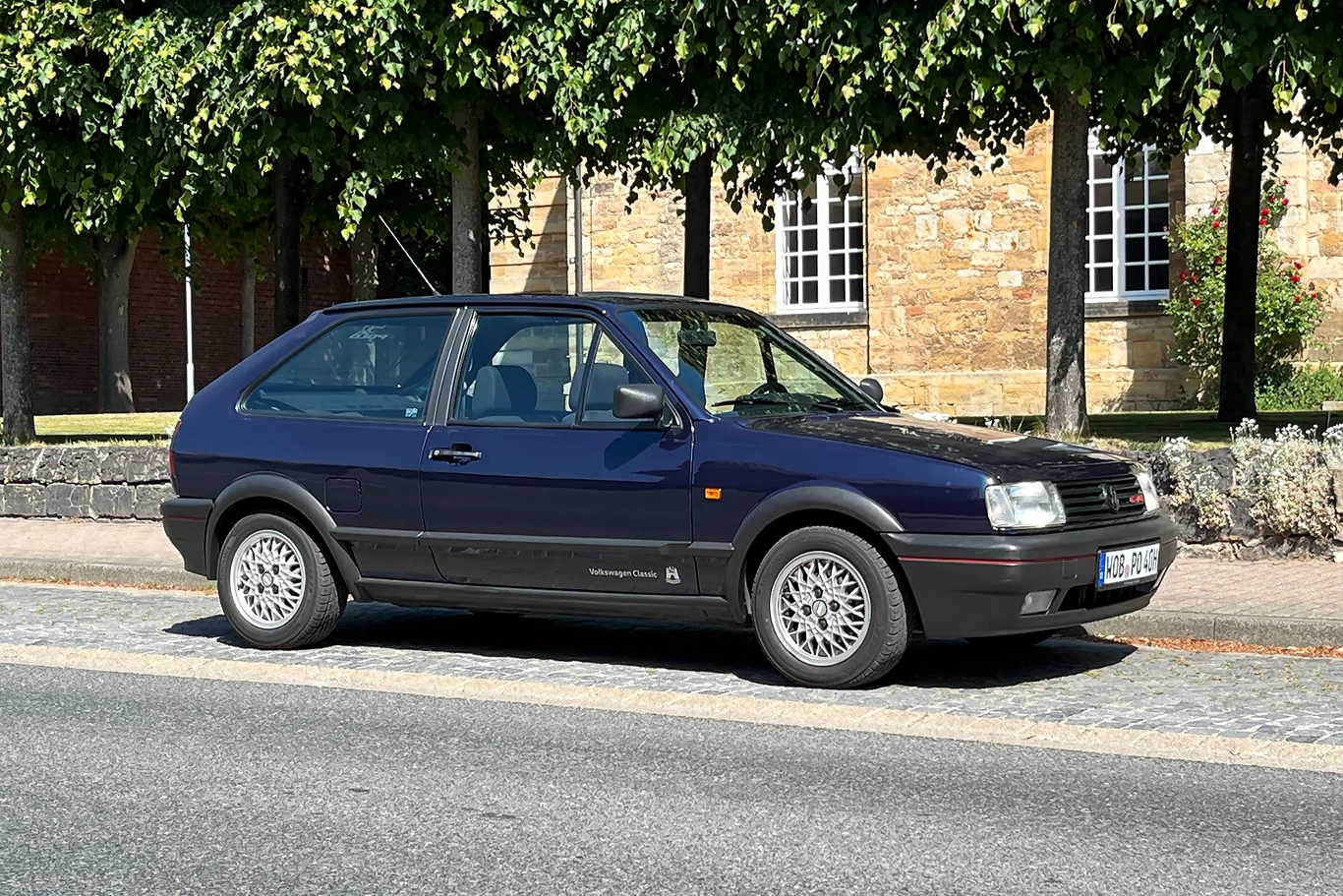 Volkswagenem Polo G40 to nie lada gratka dla miłośników klasycznych hot-hatchy. Czarny samochód osobowy marki Volkswagen Golf II zaparkowany na ulicy. W tle widoczne są drzewa oraz ceglane budynki z dużymi oknami.
