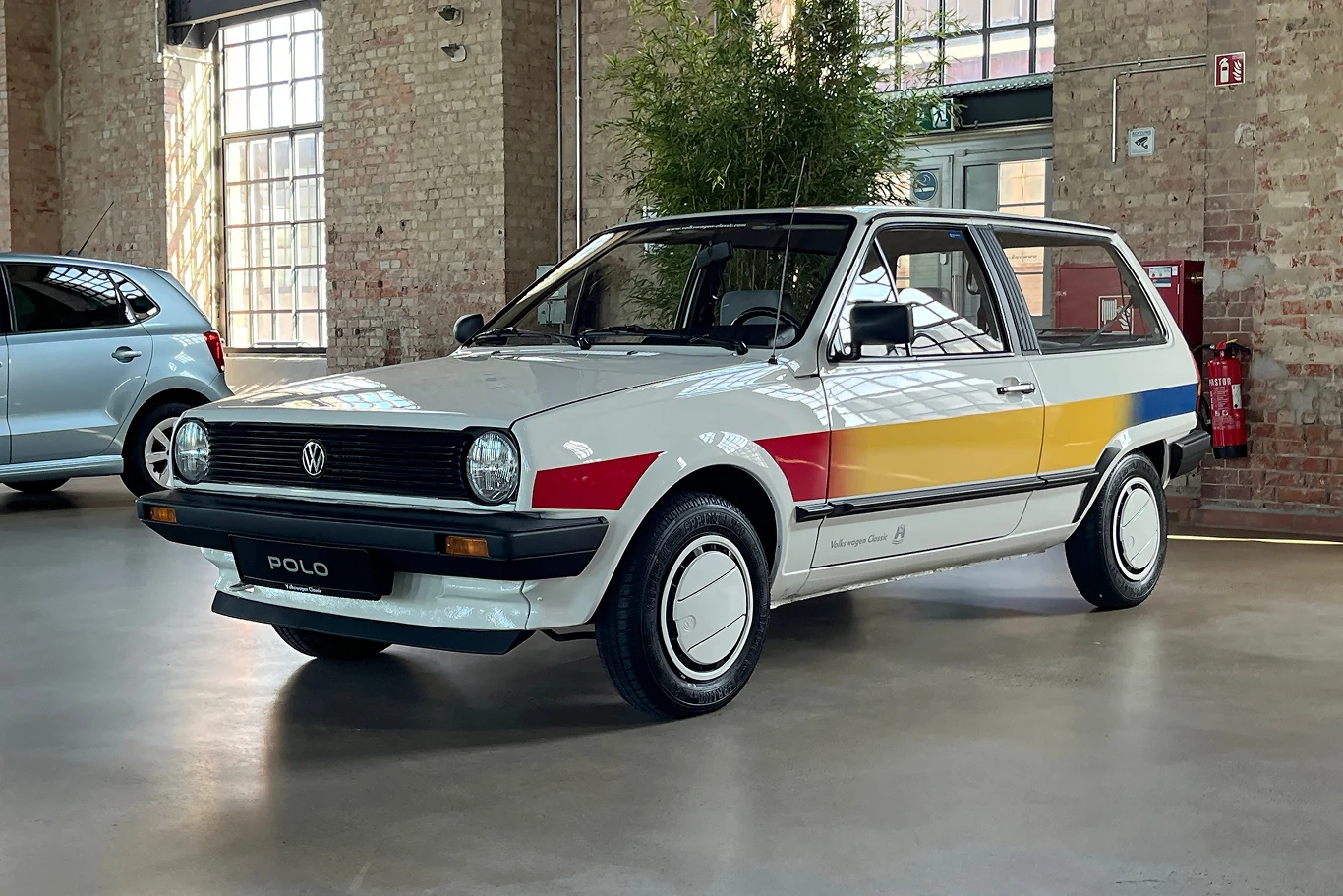 Polo II Öko-Polo spalał tylko 1,7 l oleju napędowego na 100 km. Klasyczny samochód Volkswagen Polo w białym kolorze z kolorowymi pasami na bokach, stoi w przestronnym, industrialnym wnętrzu na tle ceglanej ściany.
