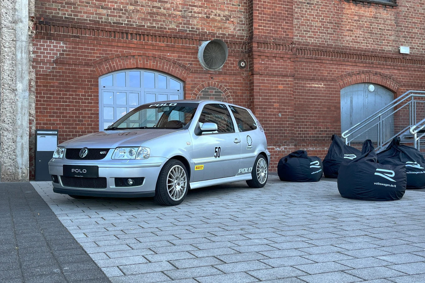 W 1999 roku zaprezentowano produkcyjną odmianę GTI wyposażoną w 1.6 l silnik o mocy 125 KM. Srebrny samochód Volkswagen Polo zaparkowany na placu przed budynkiem z cegły, obok kilka czarnych puf z nadrukiem 'R'.