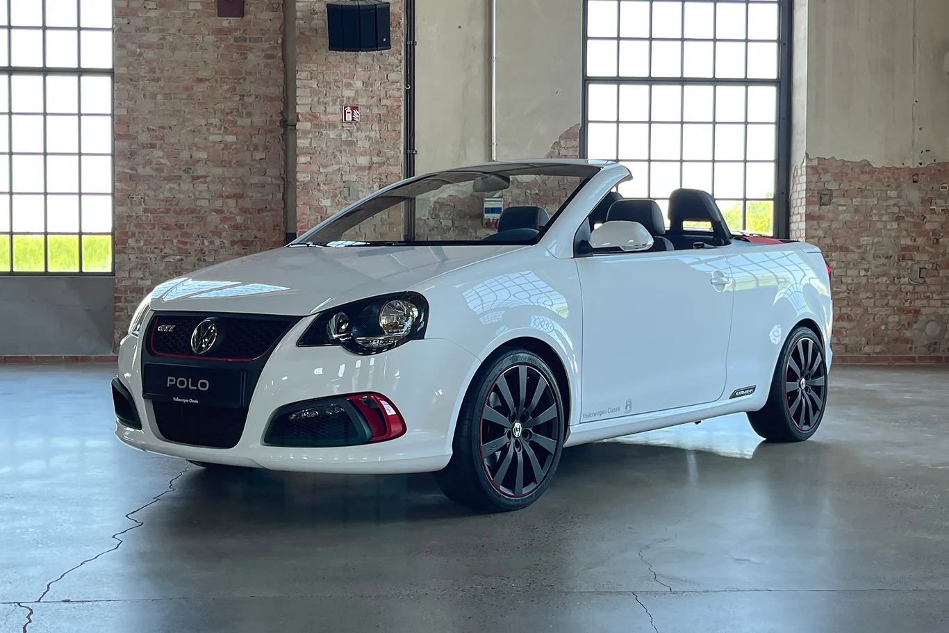 Prototyp GTI Cabrio od Karmanna nigdy nie trafiło do produkcji. Szkoda Biały kabriolet Volkswagen Polo GTI w pomieszczeniu przemysłowym z ceglaną ścianą w tle. Samochód stoi na betonowej podłodze, podkreślając nowoczesny design i sportowy charakter pojazdu.