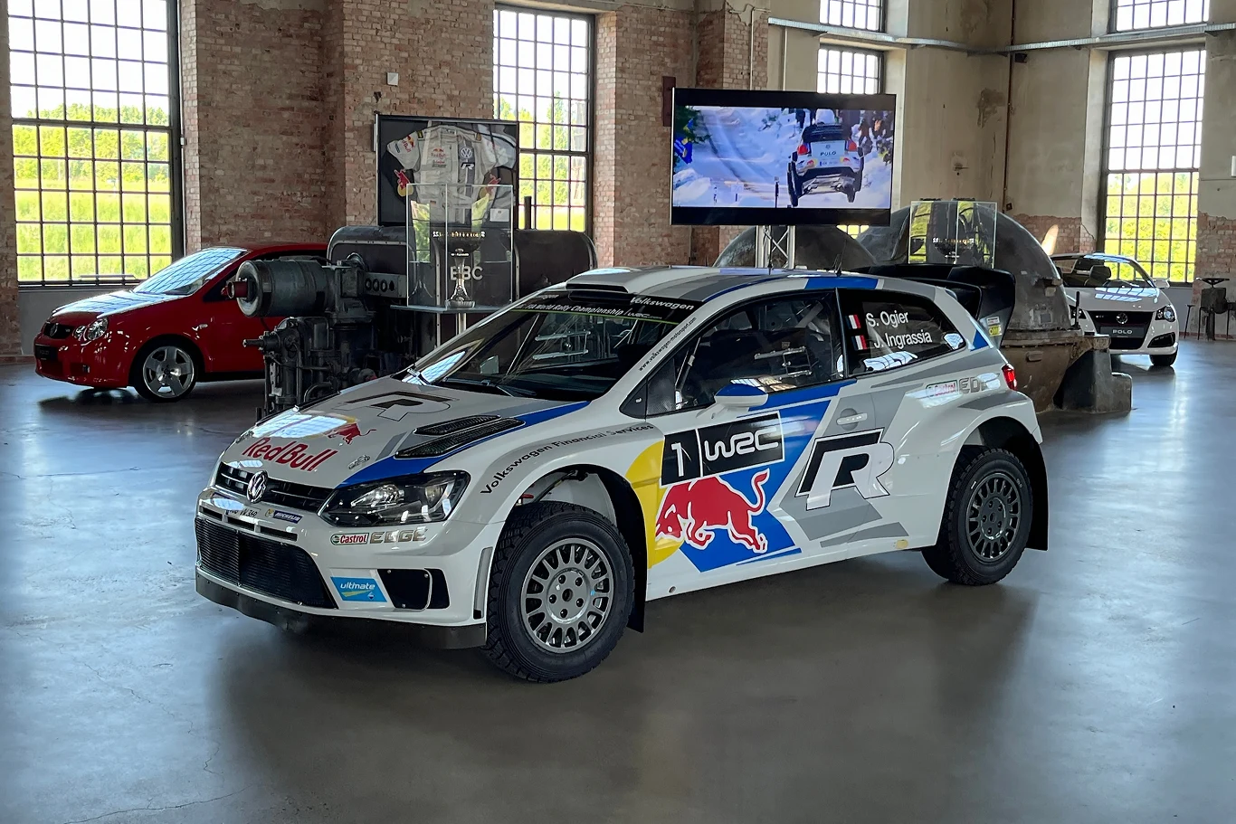 Na wiele lat Volkswagen zdominował klasyfikację WRC. Na zdjęciu Polo WRC Sebastiena Ogiera. Samochód wyścigowy Volkswagen Polo R WRC w muzeum motoryzacji, otoczony innymi pojazdami. Auto prezentuje logotypy sponsorów oraz numer rajdowy, stojąc na błyszczącej posadzce w przestronnej hali z dużymi oknami.