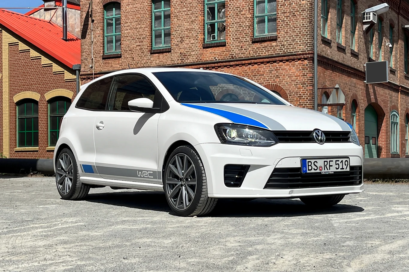 Volkswagen Polo R WRC to najmocniejsze Polo w historii modelu. Biały samochód marki Volkswagen stoi zaparkowany na placu przed budynkiem z czerwonej cegły. Samochód ma nowoczesny design z niebieskimi akcentami i sportowymi felgami.