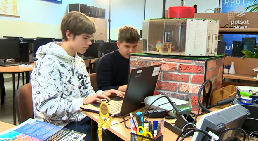 Dwóch młodych mężczyzn siedzi przy biurku w szkolnej pracowni komputerowej, skupionych na pracy przy laptopach. Przed nimi znajduje się model symulujący budynek, z elementami wykonanymi z czerwonych cegieł i szklanej obudowy.