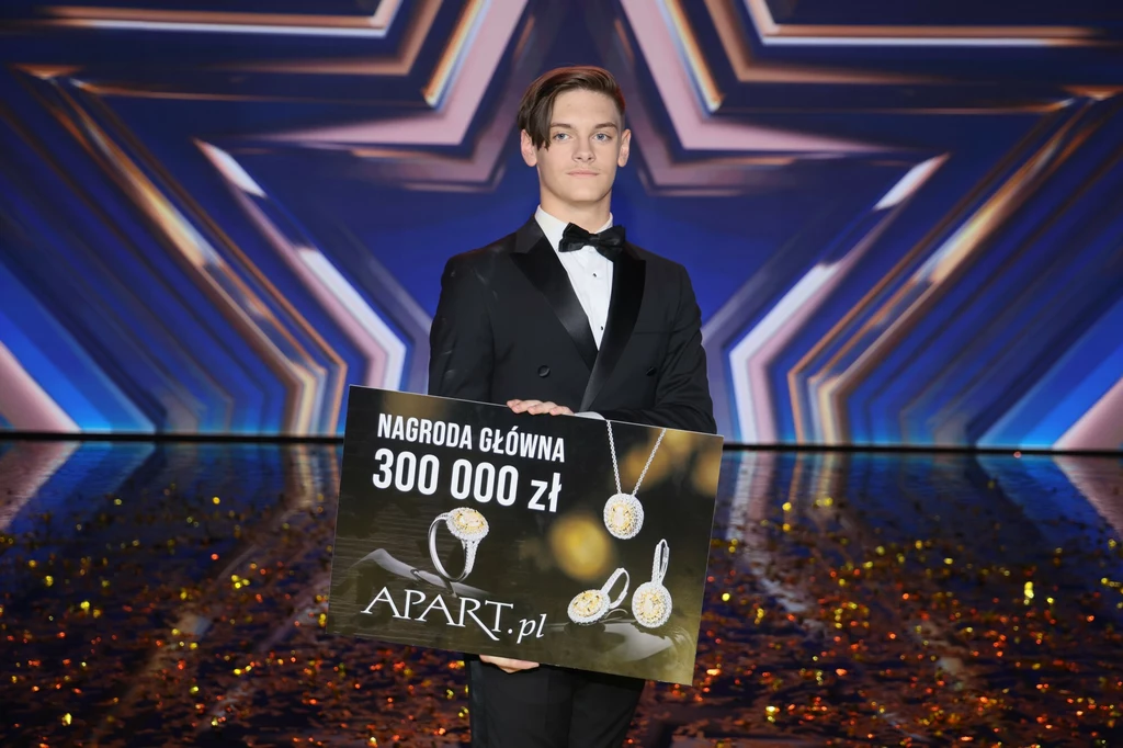 Krystian Leśnik wygrał 16. edycję "Mam talent" Młody mężczyzna w eleganckim, czarnym garniturze z muszką trzyma dużą tablicę z napisem 'Nagroda główna 300 000 zł'. Tablica zawiera również grafiki biżuterii i logo firmy Apart. Tło jest futurystycznie graficzne, z motywem gwiazdy.
