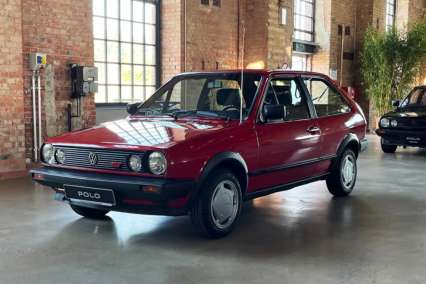 W momencie debiutu Polo G40 było szybsze od Golfa GTI i niemal dorównywało osiągami wersji GTI 16V. Klasyczny czerwony Volkswagen Polo zaparkowany w jasnym, przestronnym pomieszczeniu z ceglaną ścianą i dużymi oknami.