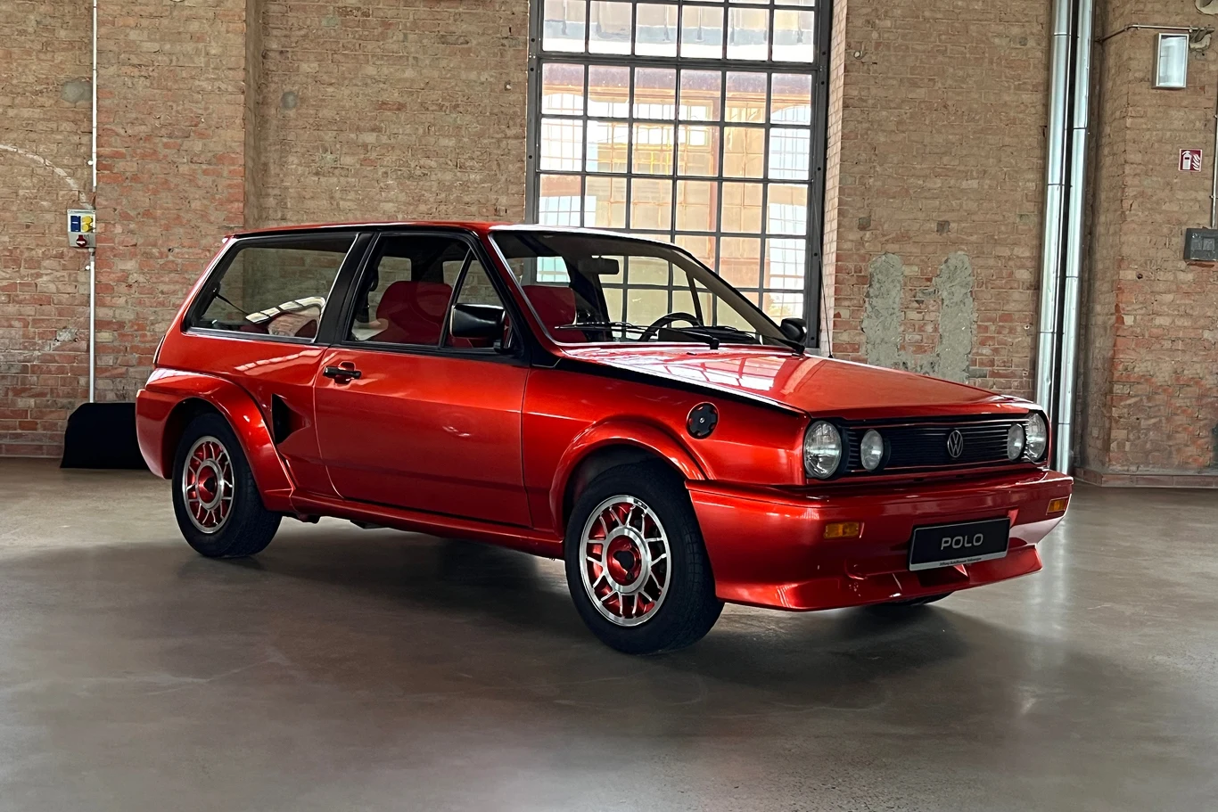 Polo zyskało wielkie uznanie wśród fanów przeróbek. Na zdjęciu jedyny w swoim rodzaju model z silnikiem typu boxer umieszczonym z tyłu pojazdu. Czerwony, klasyczny samochód Volkswagen Polo w dobrze oświetlonym warsztacie. Auto ma sportowe felgi i lśniący lakier, co przyciąga uwagę. W tle ceglane ściany i duże okno wpuszczające dużo światła.