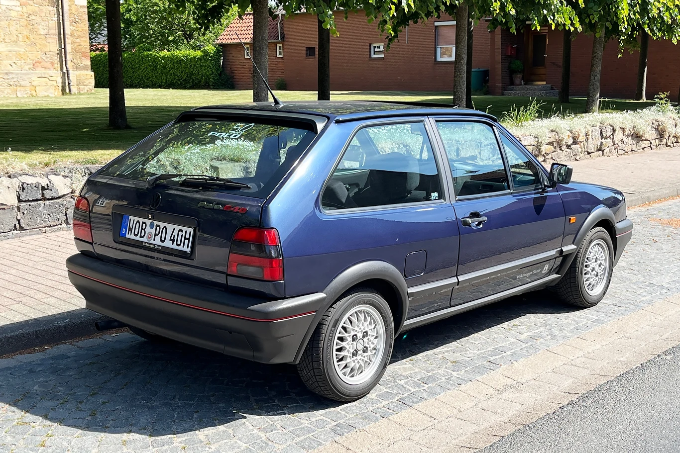 Polo G40 – również z 1,3-litrowym silnikiem wspomaganym kompresorem – ostatecznie trafiło na stałe do oferty. Samochód osobowy zaparkowany przy krawężniku, w tle ogrodzenie z kamienia oraz zielone trawniki i drzewa, które tworzą spokojne tło miejskiego krajobrazu.