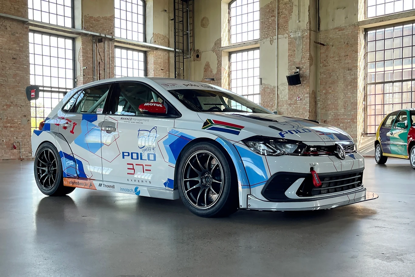 Motorsport w wydaniu Polo, to nie tylko WRC. Na zdjęciu model GTI CUP w specyfikacji wyścigowej. Samochód wyścigowy Volkswagen Polo GTI zaparkowany w przestronnej hali o surowych, ceglastych ścianach. Auto ma sportowy design i ozdobione jest licznymi naklejkami oraz logotypami sponsorów.