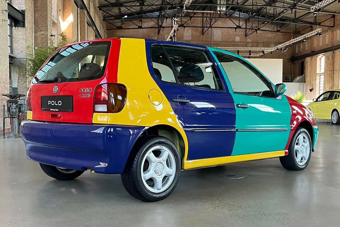 Polo Harlequin nawiązywało do reklamy Garbusa z 1964 roku, gdzie także pojawiało się auto z elementami w różnych kolorach. Samochód Volkswagen Polo w wersji harlekin, charakteryzujący się różnokolorowymi panelami karoserii, stoi w przestronnym, nowoczesnym wnętrzu z metalowymi konstrukcjami i dużymi oknami.