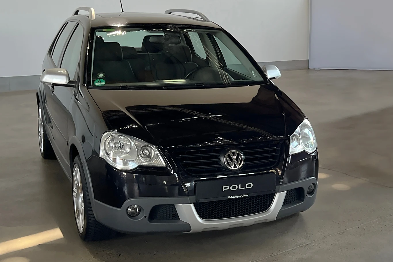 Uterenowione Polo Cross to jeden z pierwszych crossoverów producenta. Miał m.in. o 7 cm wyższy prześwit od klasycznego wariantu. Czarny samochód osobowy marki Volkswagen Polo stoi w przestronnym, jasnym pomieszczeniu. Samochód jest skierowany przodem do widza. Na tle widnieje biała ściana, a na podłodze widoczne są cienie rzucane przez auto.